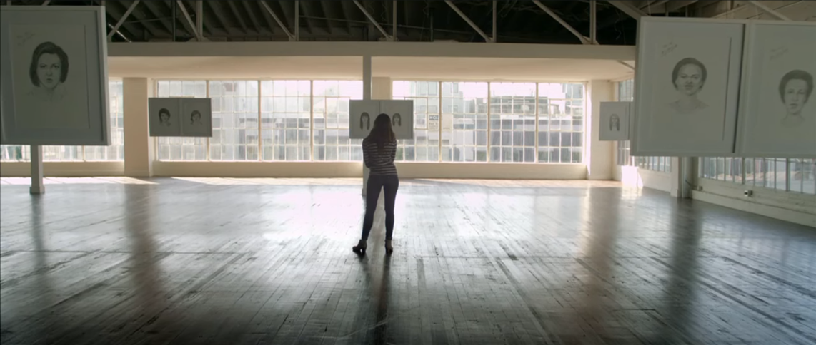 An image from the video wherein a woman is standing with many face sketches around